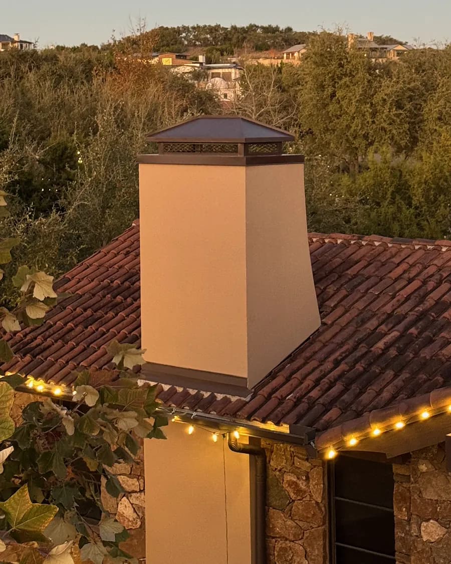A stucco chimney topped with a bronze chimney cap on a tile roof at dusk