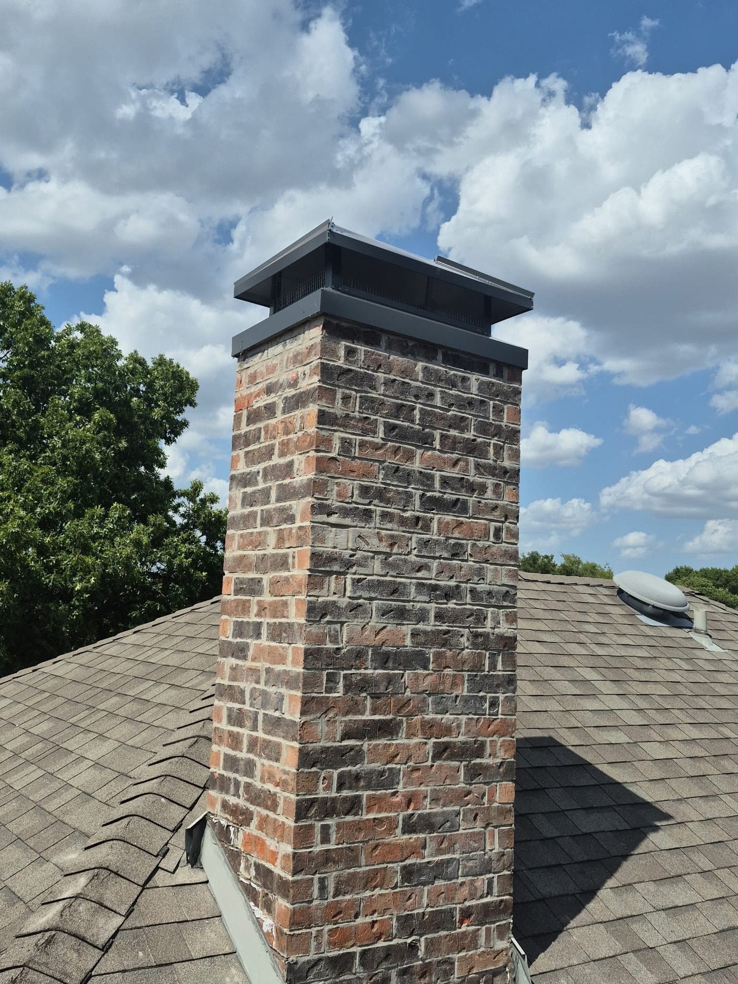 Base of a brick chimney meeting gray asphalt shingles with metal flashing along the joint
