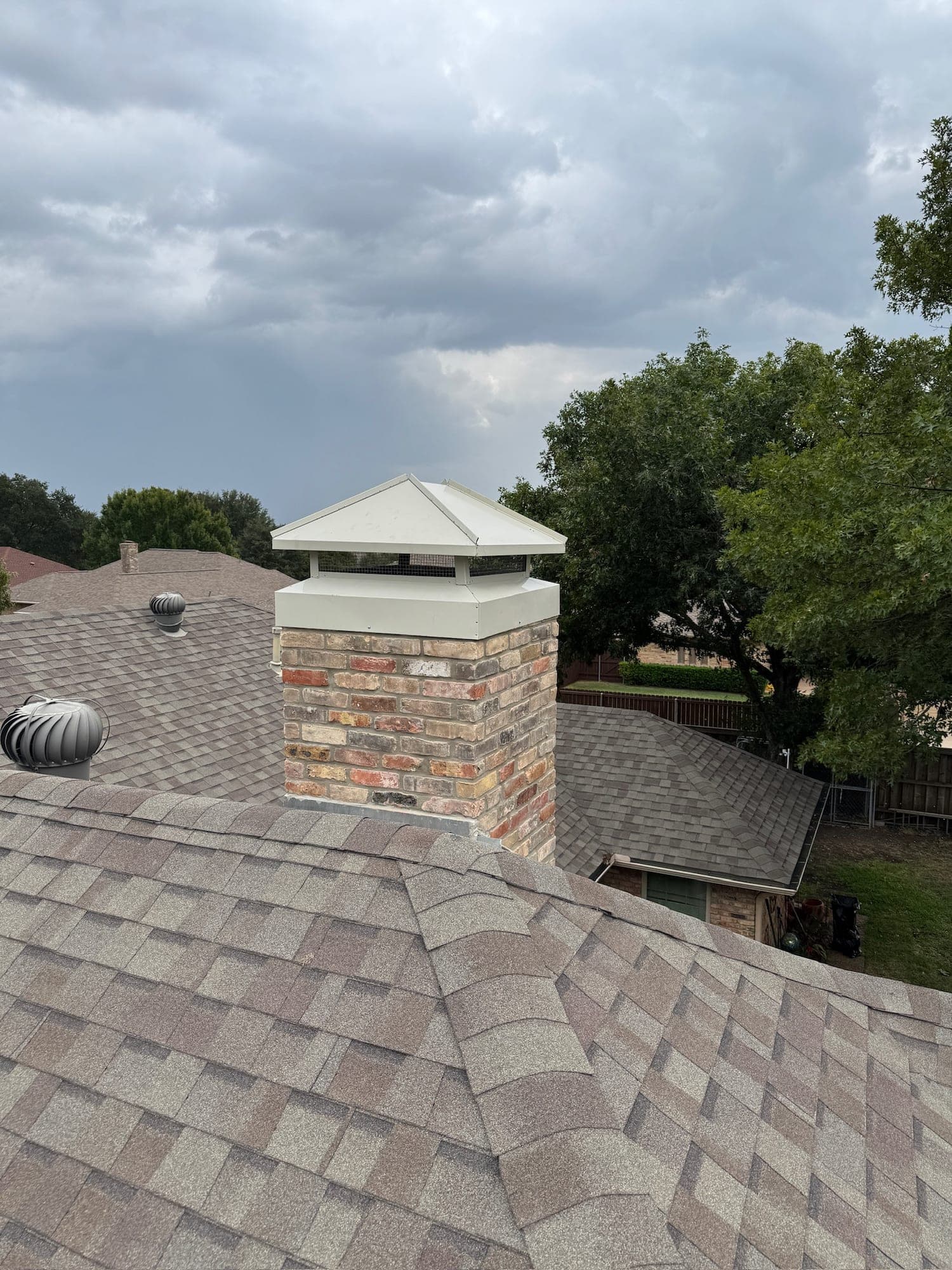 Brick chimney on a shingled Dallas roof with a white hip style chimney cap and mesh screen over the flue