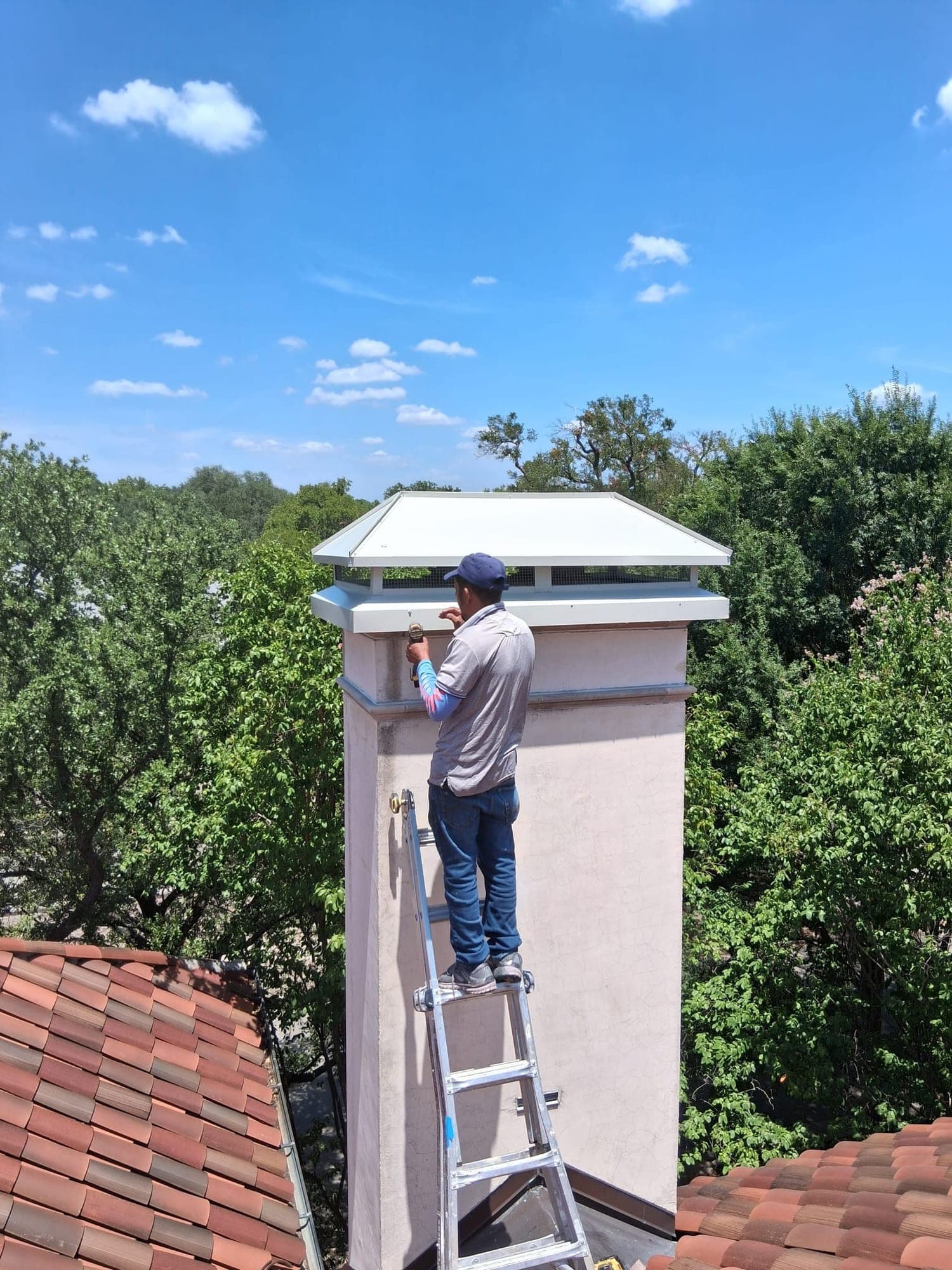 Technician on a ladder examining a large metal cap atop a tall stucco chimney above tiled rooftops